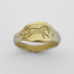 Peut inclure: Une bague dorée avec un anneau texturé et organique et une face plate légèrement irrégulière. Le centre de la bague présente un motif gravé représentant un cheval au galop entouré de petites étoiles à quatre branches de chaque côté.