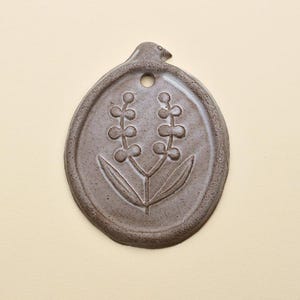 Op de afbeelding: Een bruine keramische wandhanger met een ovale vorm en een bloemenontwerp. De hanger heeft een kleine vogelachtige vorm aan de bovenkant en een gat om op te hangen. Het bloemenontwerp heeft twee stelen met kleine ronde knoppen en twee bladeren.