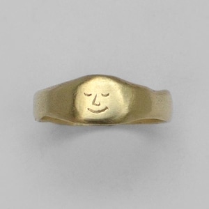 Puede incluir: Un anillo de tono dorado con una banda lisa y redondeada y una cara circular plana ligeramente elevada. El centro de la cara presenta un dibujo lineal simple y grabado de un rostro con los ojos cerrados y una pequeña sonrisa curva, creando una expresión serena.