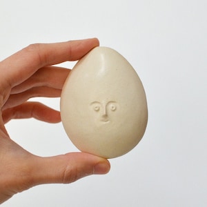 May include: A white ceramic egg with a simple face carved into it.