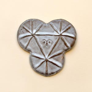 May include: A ceramic wall hanging with a distinctive design. The light grey piece features a face-like design with geometric patterns. The artwork is set against a cream-coloured background, creating a contrast that highlights the sculpture's form.