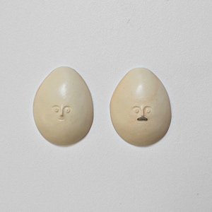 May include: Two white ceramic eggs with faces carved into them. One egg has a simple face with eyes and a mouth, while the other egg has a moustache.