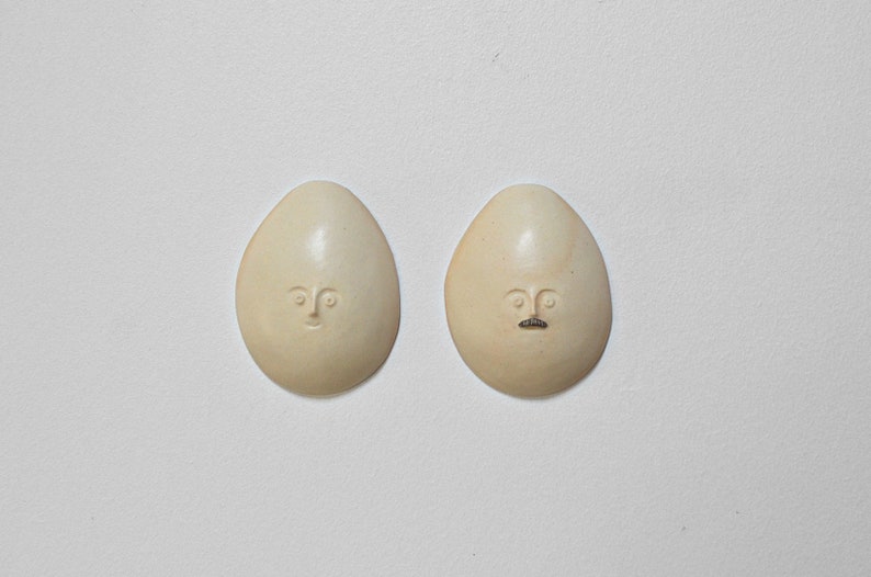 May include: Two white ceramic eggs with faces carved into them. One egg has a simple face with eyes and a mouth, while the other egg has a moustache.