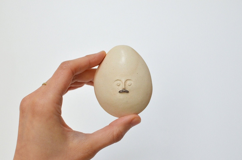 May include: A white ceramic egg with a hand-painted face, featuring eyes, a nose, and a moustache.