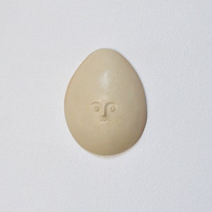 May include: A ceramic egg-shaped ornament with a simple face carved into it. The ornament is a light beige colour.