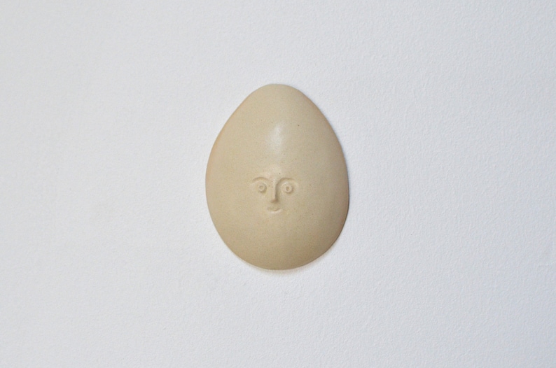 May include: A ceramic egg-shaped ornament with a simple face carved into it. The ornament is a light beige colour.