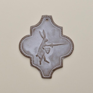 May include: A ceramic wall plaque featuring a rabbit with a sword and shield. The design is in a quatrefoil shape, with a hole for hanging. The rabbit is illustrated in a medieval style, holding a sword and shield.