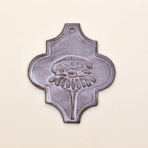 May include: A decorative, gray ceramic tile with a frog design. The frog is perched on a flower, and the tile has a unique, ornate shape. The tile is a neutral gray color and has a hole at the top for hanging.