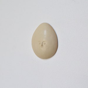 May include: A beige ceramic egg-shaped ornament with a simple face sculpted into the surface.