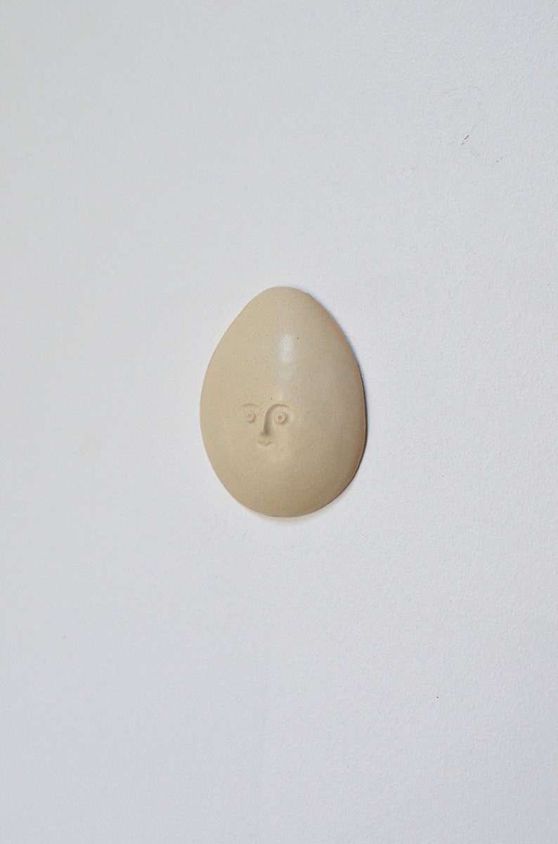 May include: A beige ceramic egg-shaped ornament with a simple face sculpted into the surface.