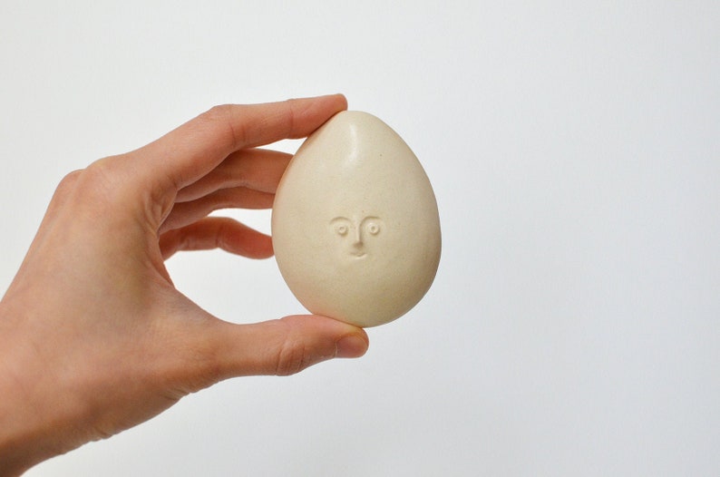 May include: A white ceramic egg with a simple face carved into it.