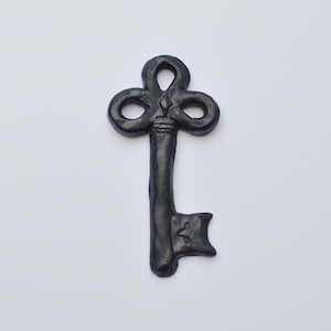 Puede incluir: Una llave de metal negro con una cabeza decorativa y adornada y una cerradura simple y plana. La llave es una pieza decorativa, no una llave funcional.
