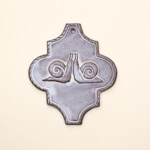 May include: A grey ceramic wall tile with a decorative design. The tile features two stylized snails facing each other. The tile has a decorative border and a hole at the top for hanging.