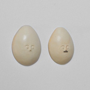May include: Two white ceramic egg-shaped ornaments with hand-painted faces. One ornament has a simple face with eyes and a mouth. The other ornament has a face with eyes, a mouth, and a moustache.
