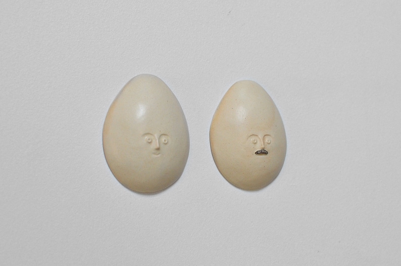 May include: Two white ceramic egg-shaped ornaments with hand-painted faces. One ornament has a simple face with eyes and a mouth. The other ornament has a face with eyes, a mouth, and a moustache.