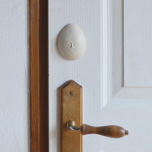 May include: A white ceramic doorstop shaped like a face with a simple design. The doorstop is attached to a white door with a wooden door handle.