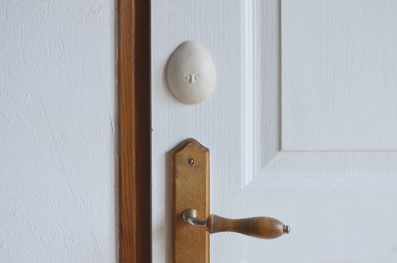 May include: A white ceramic doorstop shaped like a face with a simple design. The doorstop is attached to a white door with a wooden door handle.
