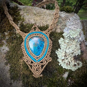 May include: A handcrafted macrame necklace featuring a large, teardrop-shaped blue and teal stone pendant. The pendant is framed with turquoise and tan macrame cord. The necklace is displayed on a moss-covered rock.
