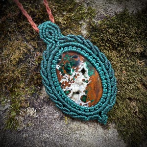 May include: A handcrafted macrame necklace featuring a large, oval-shaped stone with a mix of brown, green, and white colors. The stone is encased in teal macrame cord and suspended from a brown cord.
