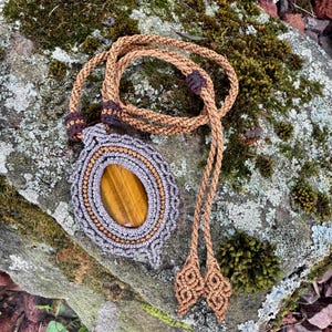 May include: A handcrafted macrame necklace featuring a large, oval tiger's eye stone pendant. The pendant is encased in gray and tan macrame, with a braided cord and decorative ends. The necklace is displayed on a moss-covered rock.