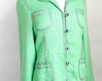 Pode incluir: Blazer verde claro com tecido texturizado. O blazer apresenta lapelas, dois bolsos frontais e acabamentos decorativos. Os botões são adornados com pedras preciosas azuis. O casaco é exibido em um manequim.