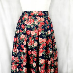 Pode incluir: Saia floral azul marinho com um padrão de rosas vermelhas e pêssego com folhas verdes. A saia tem cintura alta e um design plissado, criando uma silhueta ampla e fluida. O tecido parece ser leve.