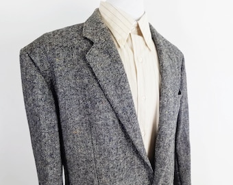 Pode incluir: Um blazer cinza-carvão e preto salpicado com tecido texturizado, apresentando lapelas entalhadas e ombros estruturados, usado sobre uma camisa de botões na cor creme claro com listras verticais finas e colarinho pontiagudo.