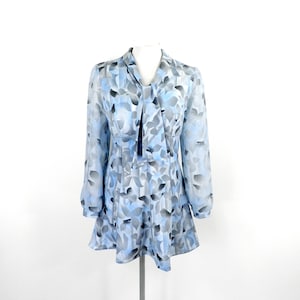 Op de afbeelding: Een lichtblauw blouse met een abstract bladmotief in grijs en wit. De blouse heeft lange mouwen en een strik bij de hals.