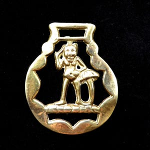 Può includere: Un ferraccio per cavalli in metallo dorato con un design a taglio di una donna in piedi su un fungo. La donna indossa una gonna e ha i capelli tirati indietro.