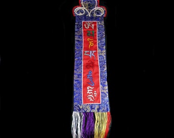 Könnte beinhalten: Ein blaues und rotes Stoffbanner mit tibetischer Schrift und einer goldenen Quaste. Das Banner hat einen dekorativen oberen Teil mit goldener Verzierung.