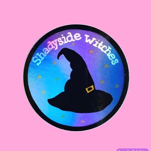 May include: A round sticker with a holographic background in shades of blue and purple. The sticker features a black silhouette of a witch's hat with a gold buckle and the text "Shadyside Witches" in white.