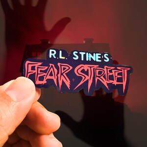 May include: A die-cut sticker featuring the text R.L. STINE'S FEAR STREET in a jagged, stylized red font against a dark blue background. The sticker has a distinct, irregular shape that follows the contour of the lettering, creating a bold visual.