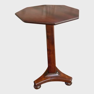 May include: A dark brown wooden pedestal table featuring an octagonal tabletop surface. The table is supported by a single central vertical column that connects to a tripod base with three curved legs, each ending in a small rounded foot.