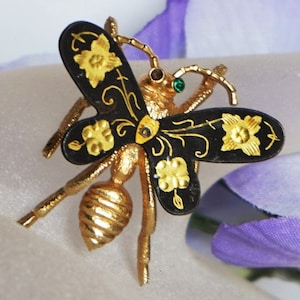 May include: A gold-toned bee brooch with black wings, adorned with gold floral designs. The bee's body is intricately detailed, and it features a small green gem. This vintage-style insect pin is a unique accessory.