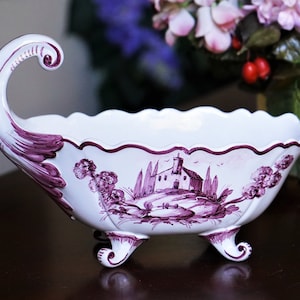 May include: White ceramic decorative bowl with a scalloped rim and three feet. The bowl features a purple landscape design with a building, trees, and floral accents. It has a curved handle on one side.