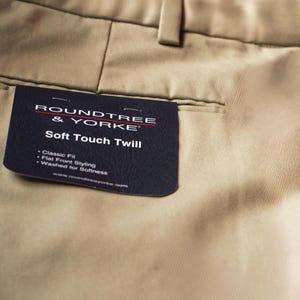 May include: Close-up of khaki-colored twill pants with a tag from Roundtree & Yorke. The tag reads "Soft Touch Twill" and lists features like "Classic Fit" and "Flat Front Styling". The pants have a classic design.
