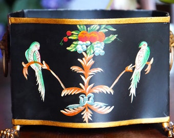 May include: Black rectangular planter with gold trim and lion's paw feet. The front features a colorful design of two parrots perched on branches, with fruit and floral accents. Decorative handles.