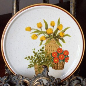 May include: A round, off-white plate with a brown rim and speckled surface. The plate features a floral design with yellow tulips, orange flowers, and green foliage in woven baskets. A decorative piece for the kitchen or dining room.