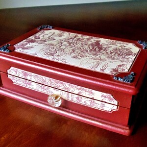 May include: A red wooden jewelry box with a decorative toile pattern on the lid and drawer. The box has a clear glass knob and ornate corner accents. The toile pattern features a detailed scene in red and white.