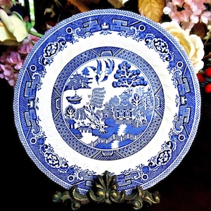 May include: A blue and white Willow pattern plate. The plate features a detailed landscape scene with a bridge, trees, and buildings. The rim has a decorative border. The plate is displayed on a decorative brass stand.