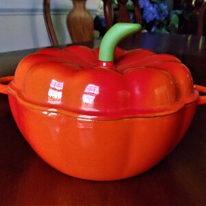 May include: A bright orange pumpkin-shaped Dutch oven with a glossy finish, featuring a light green stem-shaped handle on the lid and two side handles. The pot has a rounded, ribbed body that mimics the appearance of a real pumpkin.