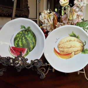 May include: Two white ceramic plates with fruit illustrations. One plate features a watermelon with a slice, and the word "watermelon". The other plate shows a melon with a slice, and the word "melon".