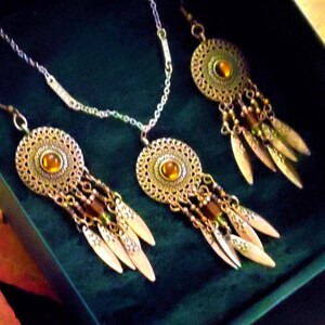 May include: A jewelry set consisting of a necklace and two matching earrings. Each piece features a circular, filigree-style metal medallion with a central amber-colored stone, accented by dangling metal leaf charms and small beads.