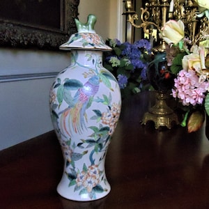 May include: A decorative, off-white ceramic vase with a lid, featuring a colorful bird and floral design. The bird has blue, pink, and yellow feathers. Green leaves and orange flowers are also painted on the vase. The lid has a green finial.