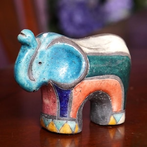 May include: A colorful ceramic elephant figurine. The elephant is painted with a turquoise head, a white back, and a body featuring orange, green, and pink sections. The legs are gray with yellow and blue triangle accents.