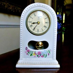 May include: A white ceramic mantel clock with a gold-tone clock face and Roman numerals. The clock has a floral design at the bottom and a gold-colored pendulum. The clock is approximately 7 inches tall.