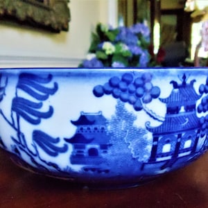 May include: A blue and white ceramic bowl with a classic Willow pattern design. The bowl features a detailed landscape scene with pagodas, trees, and a bridge. The bowl's rim is a darker shade of blue.