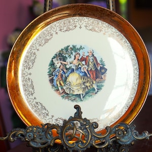 May include: A vintage decorative plate with a gold rim and a central image of a couple in period clothing. The plate has a cream-colored background with gold filigree detailing around the edge.
