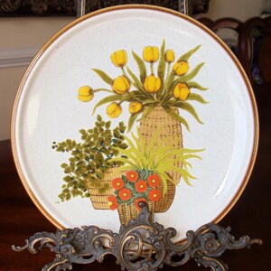 May include: A round, off-white ceramic plate with a brown rim and a floral design. The design features yellow tulips, green foliage, and orange flowers in woven baskets. The plate is displayed on a decorative metal stand.