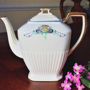 May include: White porcelain teapot with a gold handle and spout. The teapot features a decorative floral design in pink, blue, and green. The lid has a blue trim. The teapot is sitting on a dark brown surface.